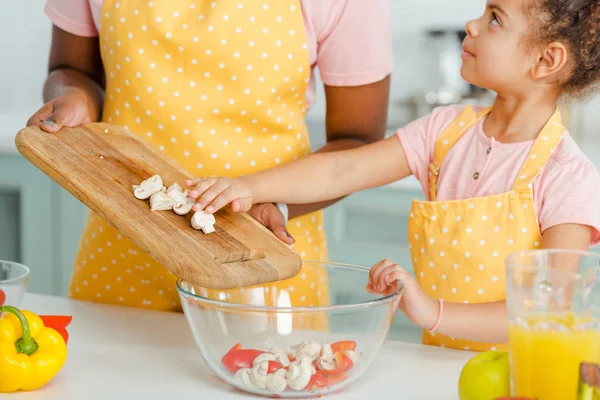 Vista recortada de la madre afroamericana sosteniendo tabla de cortar con setas cerca de cuenco y linda hija - foto de stock