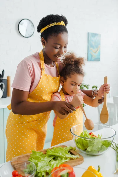 Cheerful african american mother and cute child holding wooden spoon and spatula while preparing salad — Stock Photo