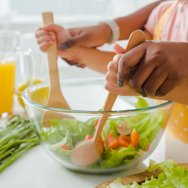 Vista recortada de la madre y el niño afroamericanos sosteniendo cuchara de madera y espátula mientras mezcla ensalada - foto de stock