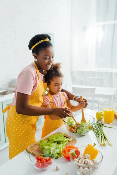 Feliz africano americano madre y hija mezcla fresco ensalada - foto de stock