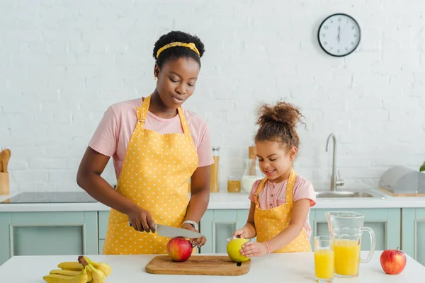 Attractive african american mother cutting apple near cute daughter — Stock Photo