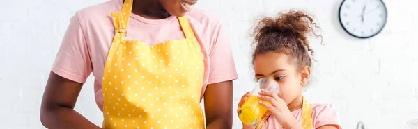 Panoramic shot of happy african american mother smiling near cute daughter drinking fresh orange juice — Stock Photo