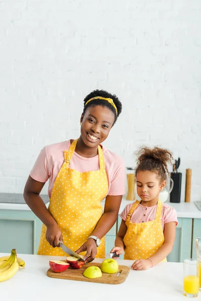 Feliz afro-americana mãe sorrindo ao cortar maçã perto da filha — Fotografia de Stock