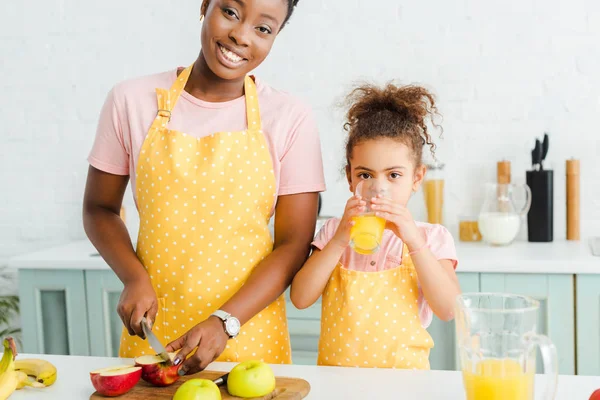 Happy african american mother smiling while cutting apple near daughter drinking orange juice — Stock Photo