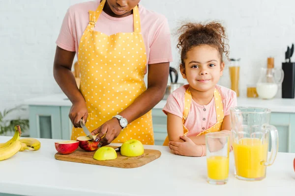 Vista ritagliata di afro americano madre taglio mela vicino carino figlia guardando la fotocamera — Foto stock