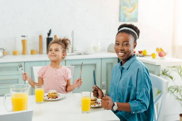 Feliz africano americano madre y hija holding cubertería cerca sabroso panqueques - foto de stock