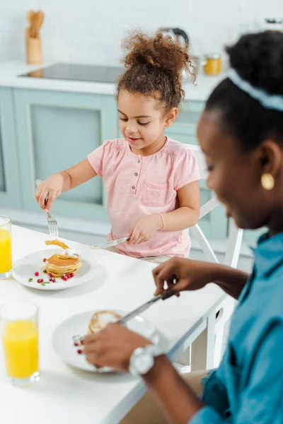 Selective focus of happy african american kid and mother holding cutlery near pancakes — Stock Photo
