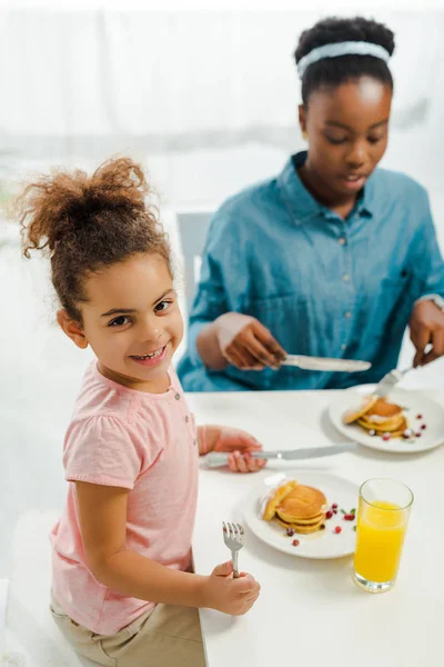 Enfoque selectivo de niño afroamericano feliz sosteniendo cubiertos cerca de panqueques y madre - foto de stock