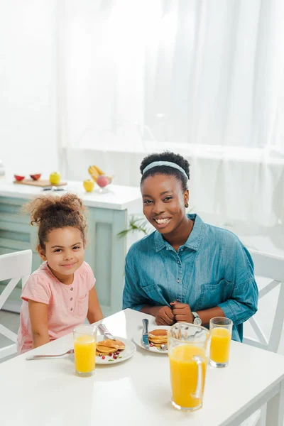 Joyeuse mère afro-américaine et sa fille regardant la caméra près de délicieuses crêpes — Photo de stock