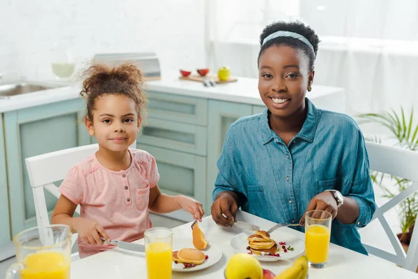 Madre e hija afroamericana sosteniendo cubiertos cerca de sabrosos panqueques en platos - foto de stock