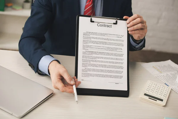 Cropped view of agent holding clipboard with contract lettering and pen near laptop and calculator — Stock Photo