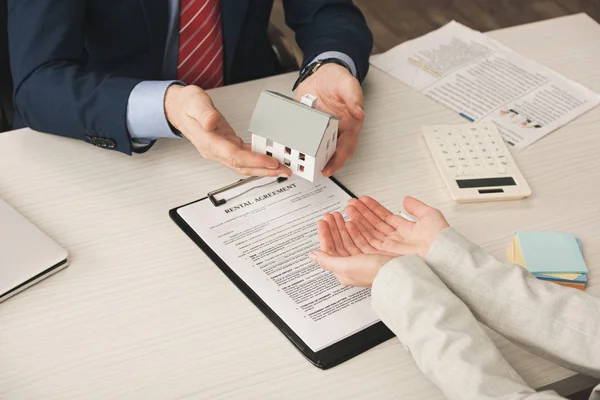 Vista recortada de agente inmobiliario dando modelo de casa a la mujer con las manos ahuecadas cerca de contrato de alquiler - foto de stock