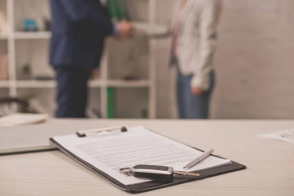 Selective focus of clipboard with document on desk near car key and two people shaking hands — Stock Photo