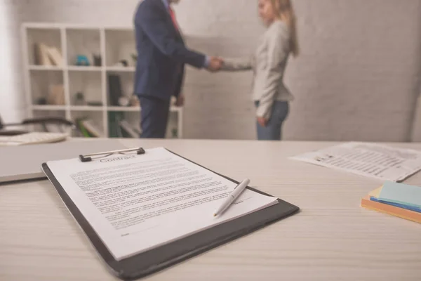 Selective focus of clipboard with document on table near agent and client shaking hands — Stock Photo