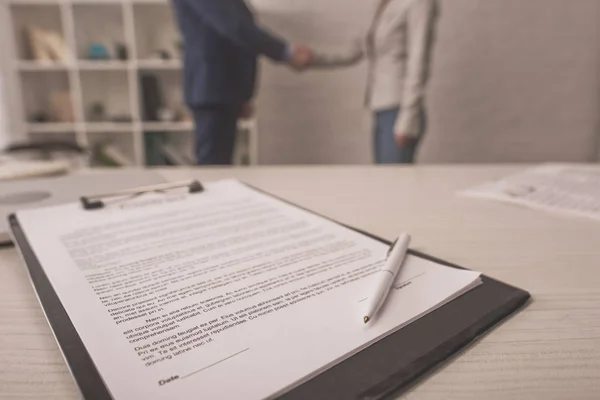 Selective focus of clipboard with document on desk near agent and client shaking hands — Stock Photo