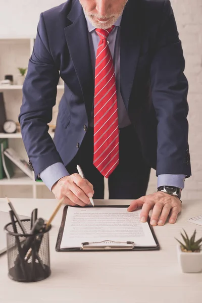 Selective focus of bearded broker signing document near stationery and plant on desk — Stock Photo