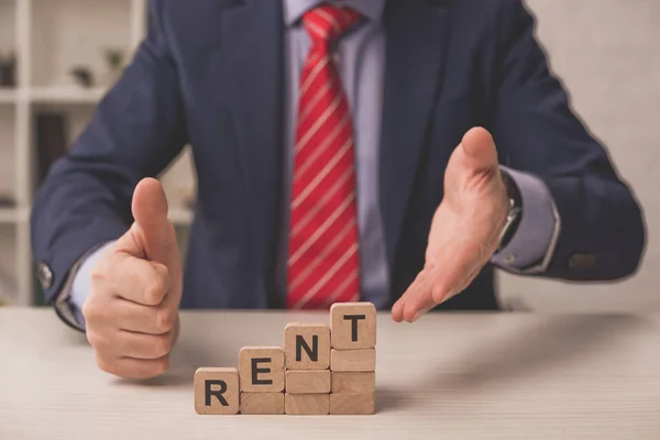 Vue recadrée de l'agent montrant pouce vers le haut et pointant à la main vers des cubes en bois avec lettrage de loyer — Photo de stock