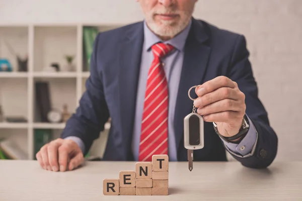 Cropped view of bearded agent holding car key near wooden cubes with rent lettering — Stock Photo