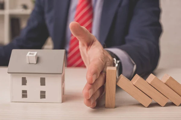 Vista recortada del agente poniendo la mano en el escritorio entre los cubos de madera y el modelo de casa de cartón - foto de stock