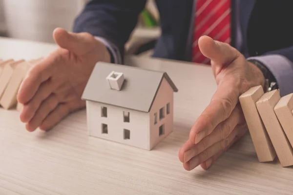 Vista recortada del agente poniendo las manos en el escritorio entre los cubos de madera y el modelo de casa de cartón — Stock Photo