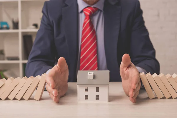 Cropped view of agent putting hands on table between wooden cubes and carton house model — Stock Photo