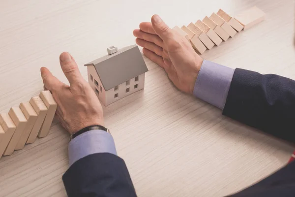 Vista recortada de agente inmobiliario poniendo las manos en la mesa entre los cubos de madera y el modelo de casa de cartón - foto de stock