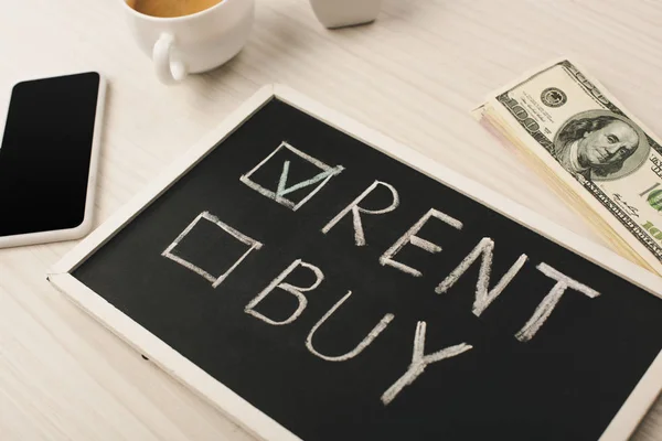 Pizarra con alquiler y comprar letras cerca de la taza de café. smartphone con pantalla en blanco y dinero - foto de stock