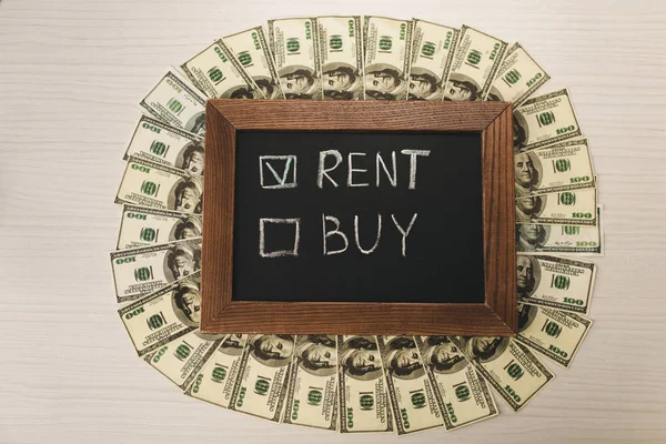 Top view of chalkboard with rent and buy lettering near dollar banknotes on desk — Stock Photo