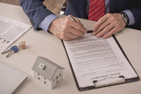 Cropped view of agent holding pen near clipboard with rental agreement, carton house model and keys on desk — Stock Photo