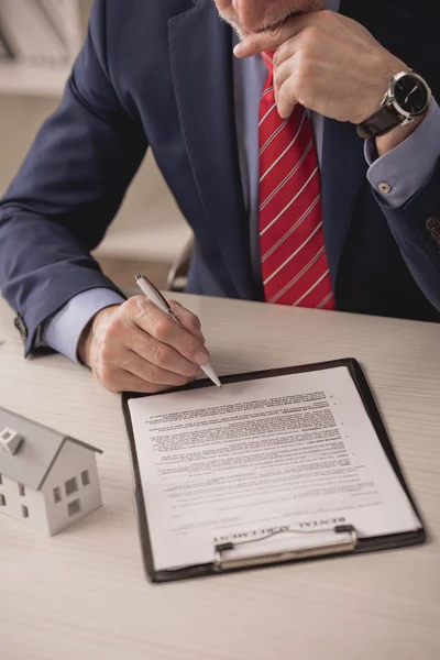 Ausgeschnittene Ansicht des nachdenklichen Agenten mit Stift in der Nähe von Klemmbrett mit Mietvertrag und Kartonhaus-Modell — Stockfoto