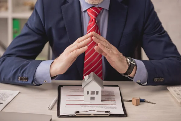 Ausgeschnittene Ansicht eines Agenten, der sich händchenhaltend über Kartonhausmodell und Klemmbrett mit Versicherungsaufdruck hält — Stockfoto