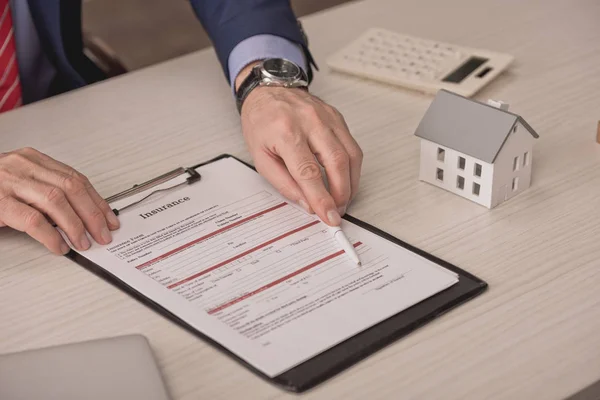 Vista recortada de la pluma de retención de agente cerca del portapapeles con letras de seguro y modelo de casa de cartón — Stock Photo