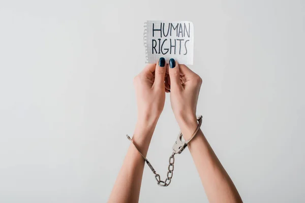 Vista recortada de una joven con esposas en las manos sosteniendo papel con letras de derechos humanos aisladas en blanco - foto de stock