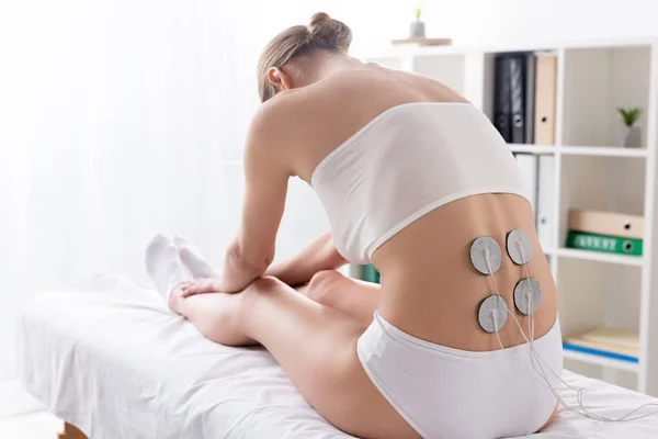 Rückansicht einer Frau in Unterwäsche, die während der Elektrodenbehandlung des Rückens in der Klinik auf der Massagecouch sitzt — Stockfoto
