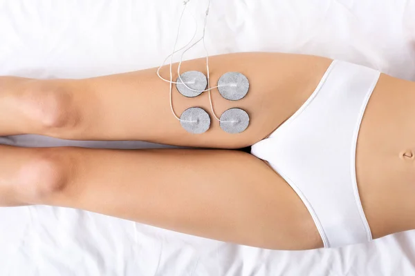 Vue du dessus de la jeune femme en culotte couchée sur le canapé de massage pendant le traitement de l'électrode de la jambe — Photo de stock