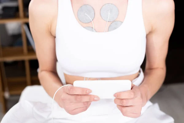 Vue recadrée de la femme tenant smartphone pendant l'électrothérapie de la poitrine en clinique — Photo de stock