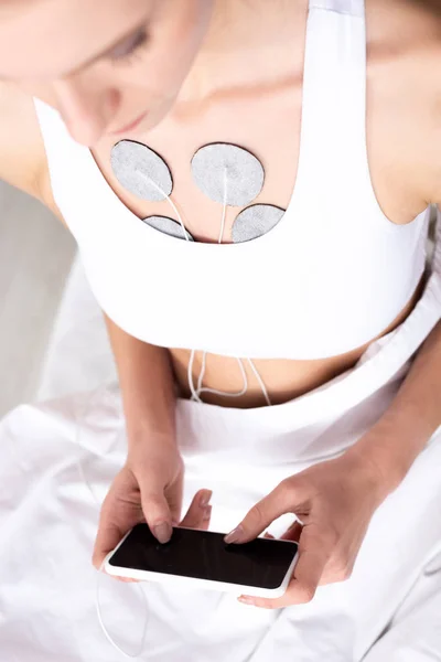 Selective focus of young woman holding smartphone during electrotherapy of chest on massage couch — Stock Photo