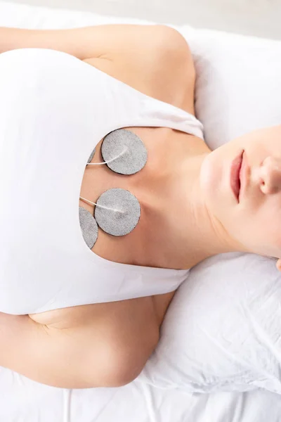 Top view of woman with electrodes on chest during electrotherapy on massage couch on grey background — Stock Photo