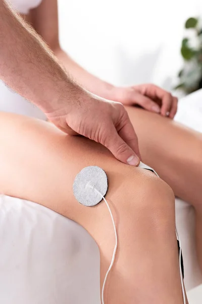 Vista recortada del electrodo de ajuste del terapeuta en la rodilla del paciente durante la electroterapia - foto de stock