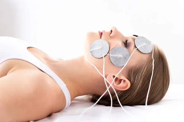 Seitenansicht der schönen Frau auf Massagecouch liegend während der Gesichtselektrotherapie auf weißem Hintergrund — Stockfoto