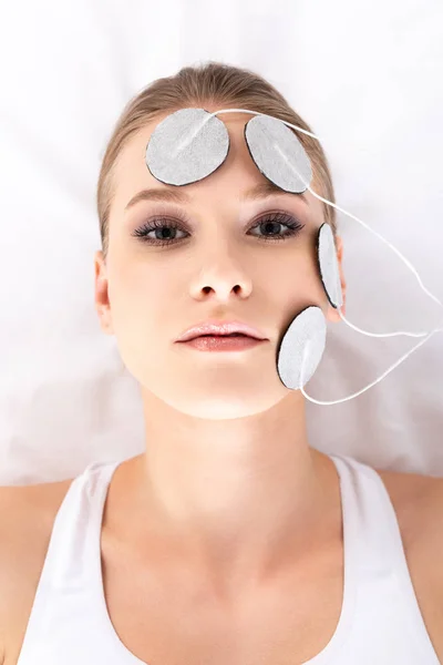 Visão superior da menina atraente olhando para a câmera durante a eletroterapia facial no sofá de massagem — Fotografia de Stock