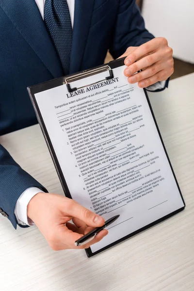 Vista recortada del agente sujetando portapapeles con letras de contrato de arrendamiento cerca de escritorio - foto de stock