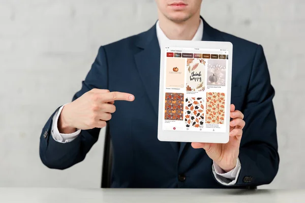 Vista recortada de agente inmobiliario en traje apuntando con el dedo a la tableta digital con aplicación pinterest en blanco - foto de stock