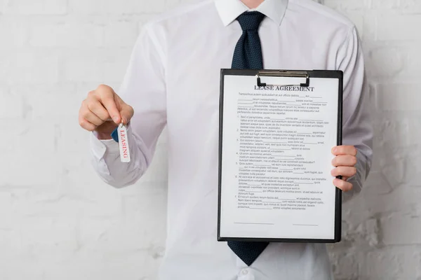 Vista recortada de agente inmobiliario en traje con llave y portapapeles con contrato de arrendamiento en blanco - foto de stock