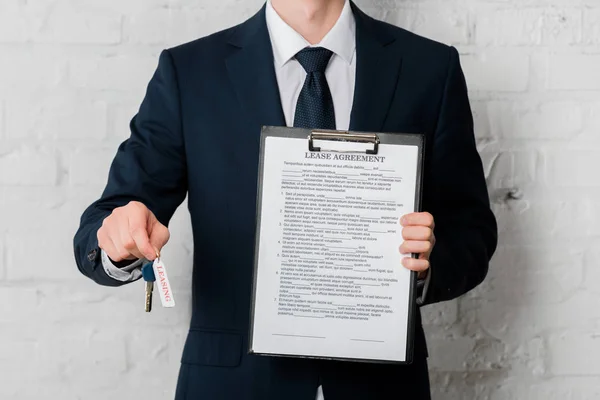 Cropped view of realtor in suit holding key with leasing lettering and clipboard with lease agreement on white — Stock Photo