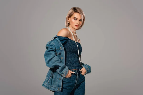 Mujer atractiva en chaqueta de mezclilla y jeans mirando a la cámara aislada en gris - foto de stock