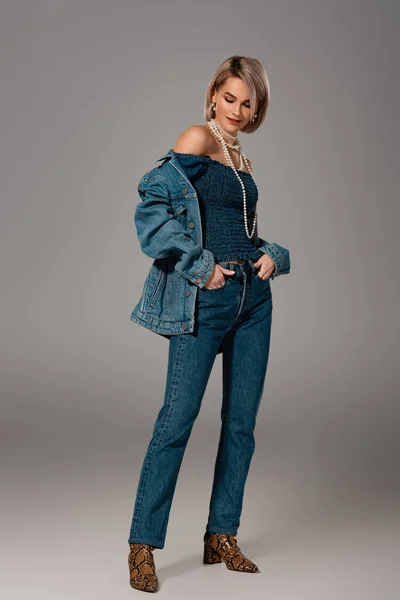 Mujer atractiva y sonriente en chaqueta de mezclilla y jeans sobre fondo gris - foto de stock