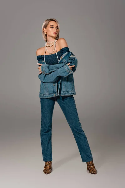 Atractiva mujer en chaqueta de mezclilla y jeans mirando hacia otro lado sobre fondo gris - foto de stock