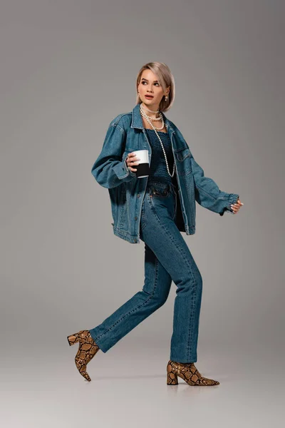 Mulher atraente em jaqueta de ganga e jeans segurando caneca isolada no fundo cinza — Fotografia de Stock