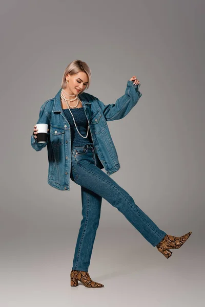 Улыбающаяся женщина в джинсовой куртке и джинсах с изолированной кружкой на сером фоне — стоковое фото